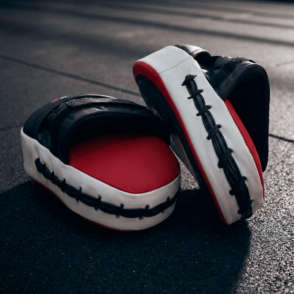 Pair of black, red, and white with black stitching Indus Cross Ventus-GL Boxing Focus Mitts one on top of one another on a dark surface.