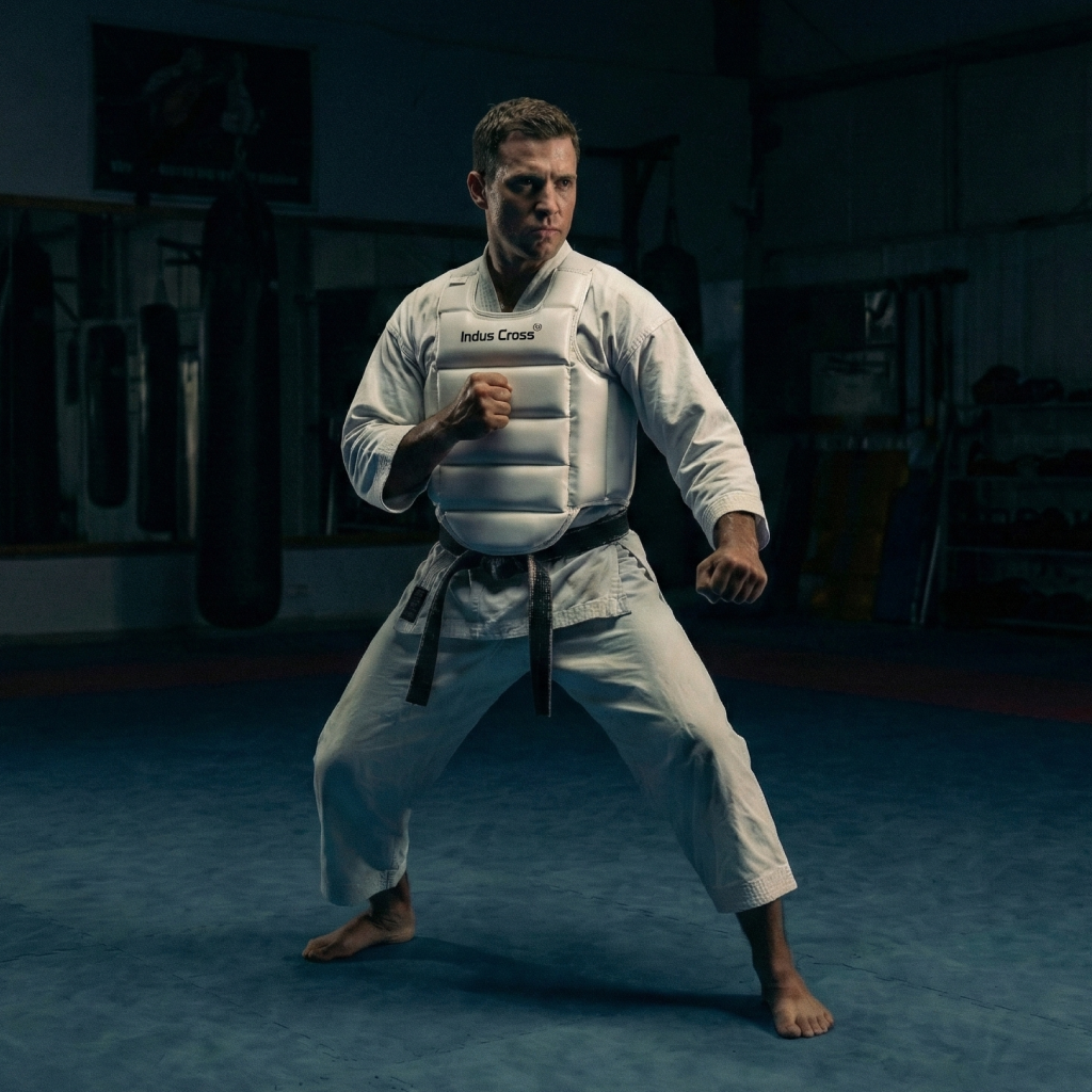 Person in a white martial arts uniform and a white karate body protector with a black belt in a dark dojo.